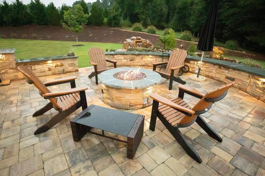 Patio scene with five wooden chairs around a circular stone fire pit, trees and landscaped garden visible in the background