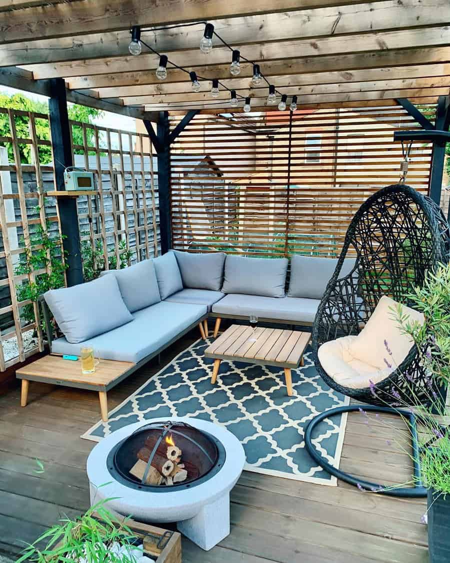 Cozy outdoor patio with gray sectional couch, hanging egg chair, wooden pergola, and fire pit on a patterned rug