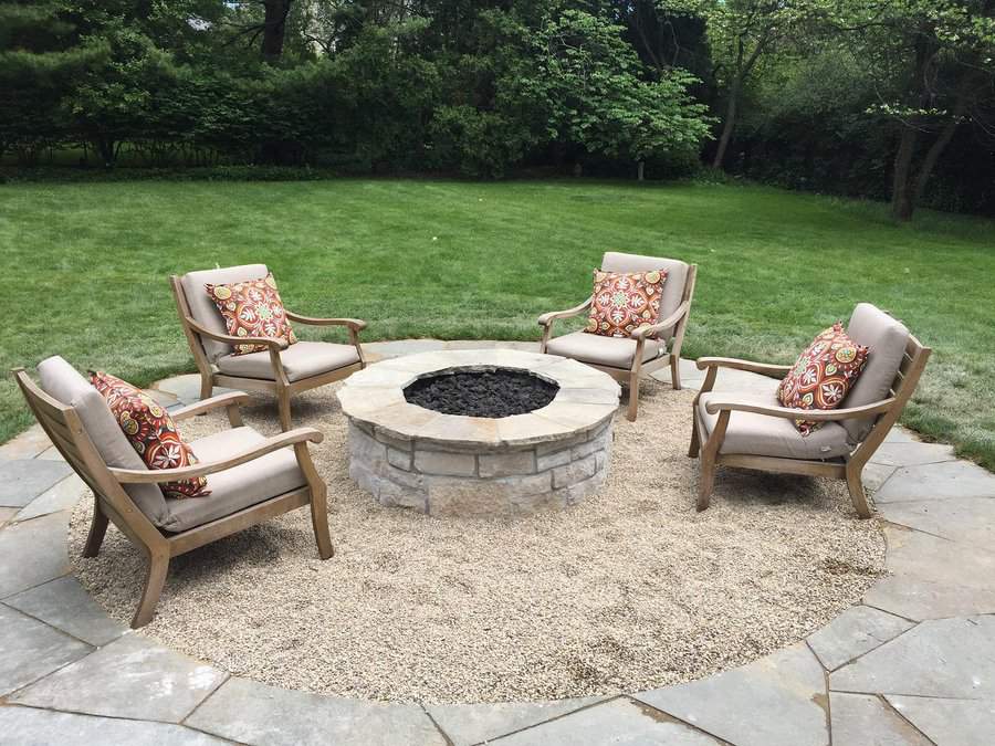 Four cushioned chairs with patterned pillows surround a stone fire pit on a gravel patio, set in a grassy backyard