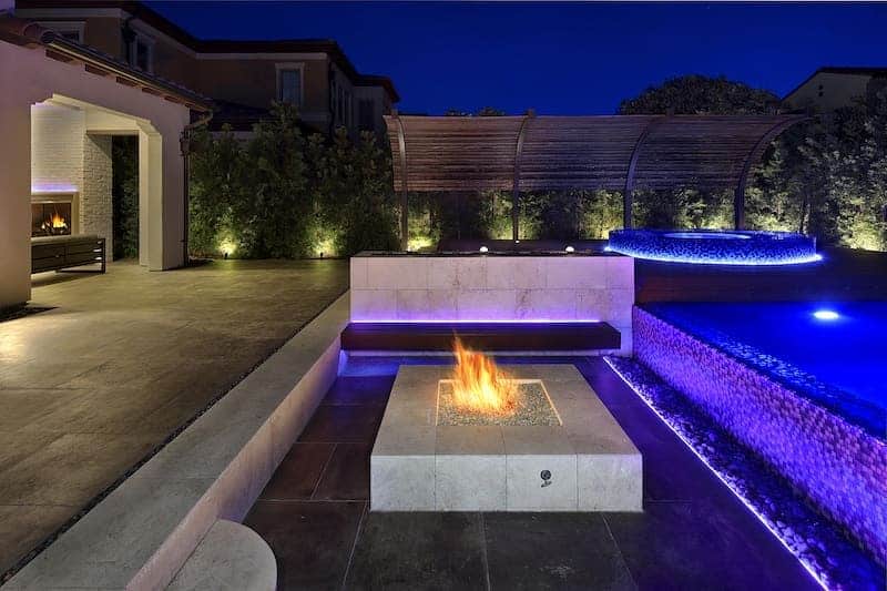 Modern patio at night with a fire pit, illuminated pool, and ambient garden lighting under a dark blue sky