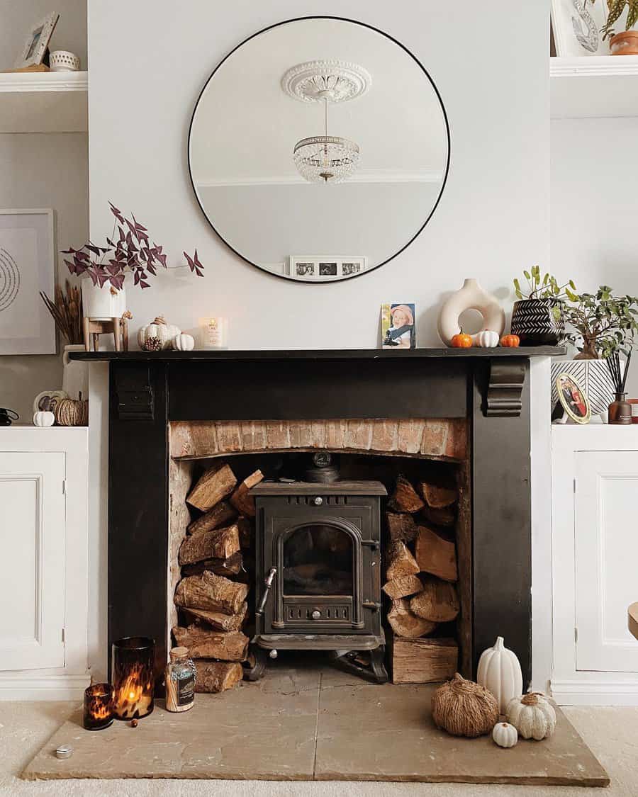 A cozy fireplace with wood logs, candles, and pumpkin decor, under a round mirror