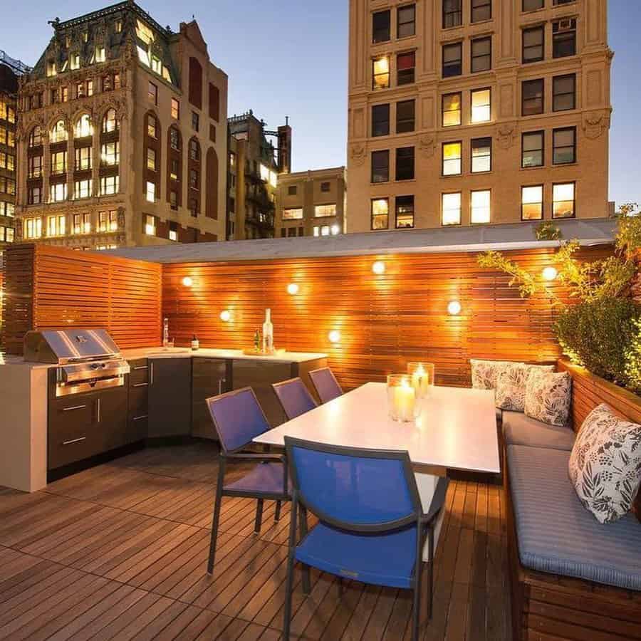 Modern rooftop terrace with wooden decking, a dining table, blue chairs, cushioned bench seating, string lights, and city views at dusk