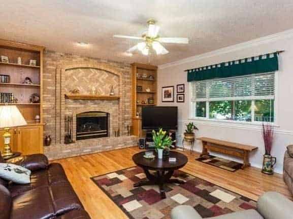 Cozy living room with fireplace, ceiling fan, TV, and a coffee table; shelves with books and plants add charm to the brick interior