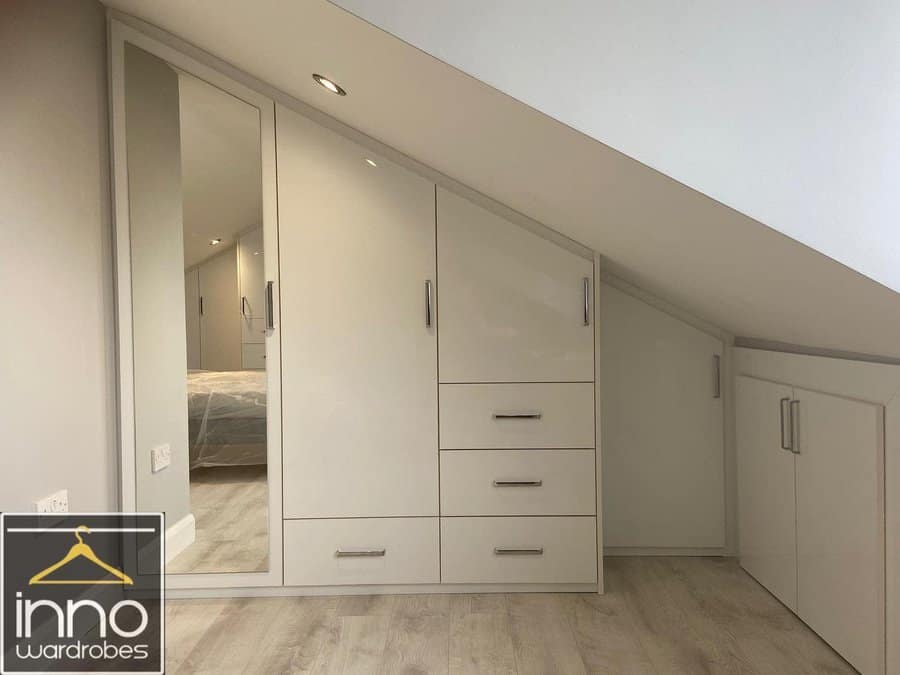 Custom-fitted attic wardrobe with sleek white cabinets, built-in drawers, and a full-length mirror, maximizing storage under a sloped ceiling.