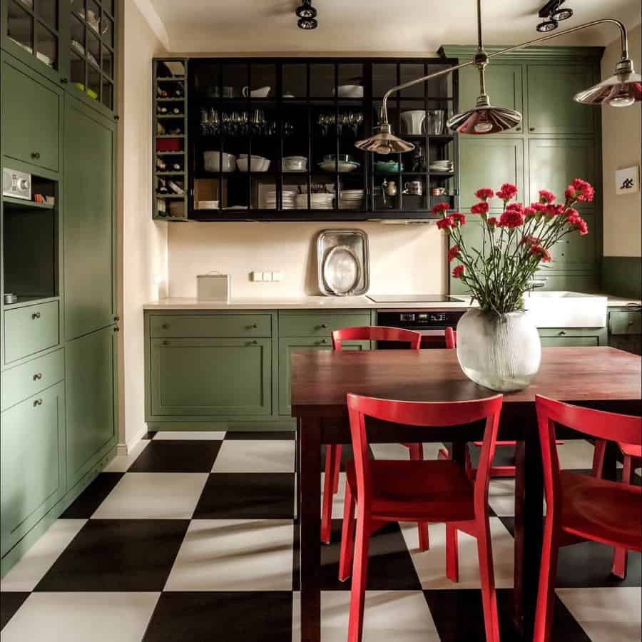 Kitchen with green cabinets, black and white checkered floor, red chairs around a dark wooden table, and red flowers in a white vase