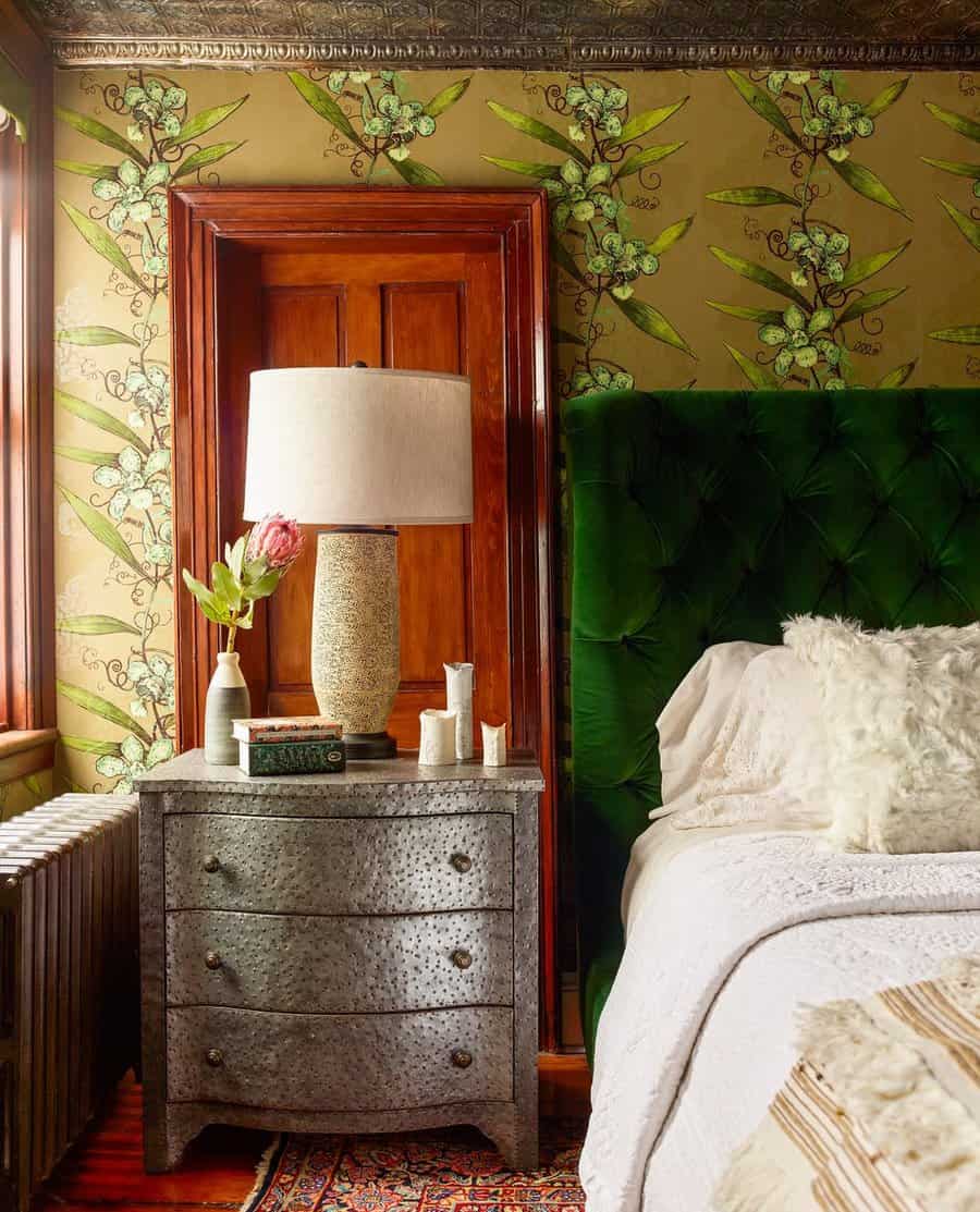 Bedroom with a green velvet headboard, floral wallpaper, and a vintage silver nightstand holding a lamp and decor items