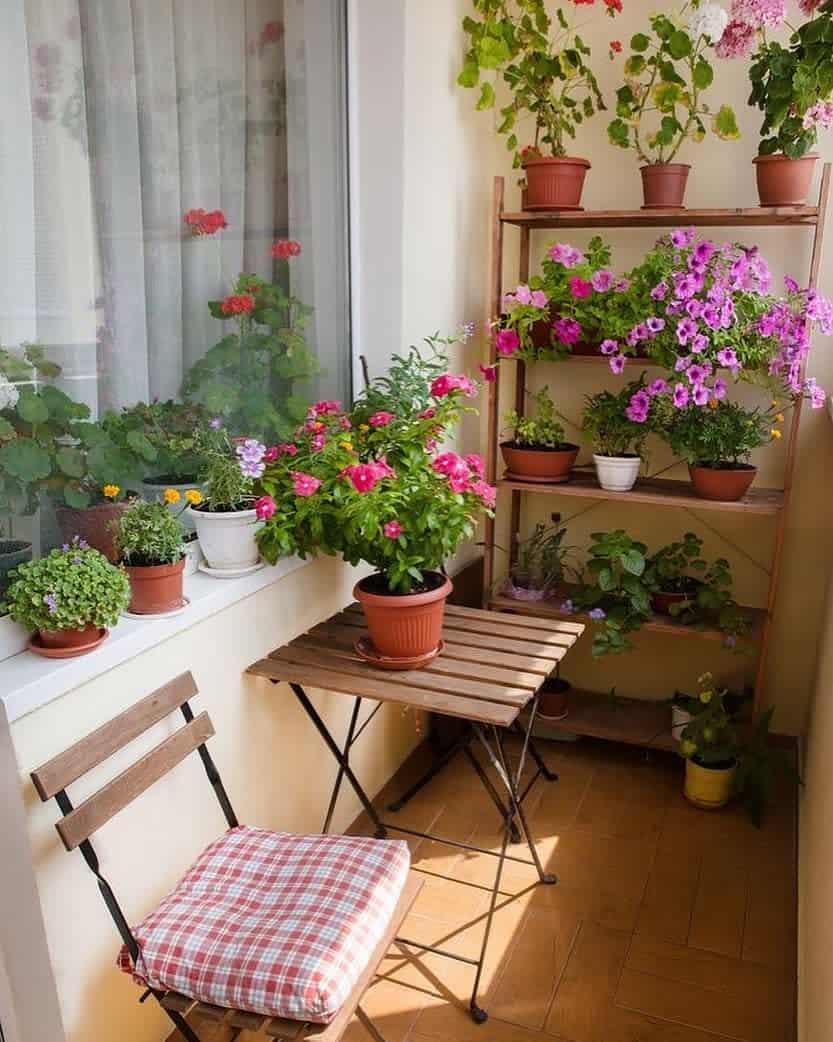 A small balcony with a wooden chair and table, decorated with various potted plants and flowers on shelves and window ledge