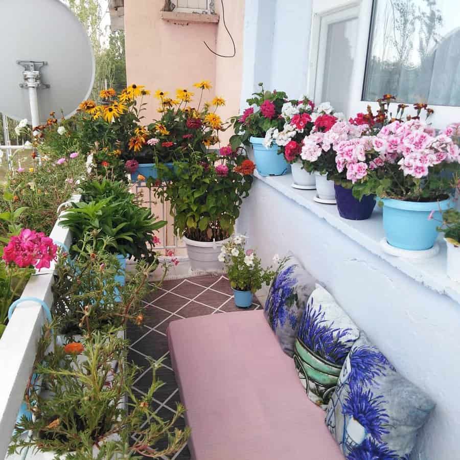 A small balcony with colorful flower pots on railings and ledges, a pink cushioned bench, and patterned pillows