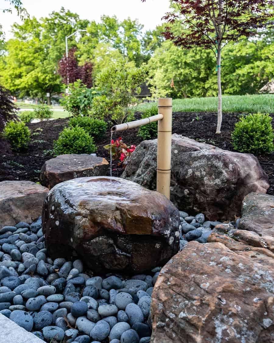 Zen garden with a bamboo water spout over rocks and pebbles, surrounded by green plants and trees