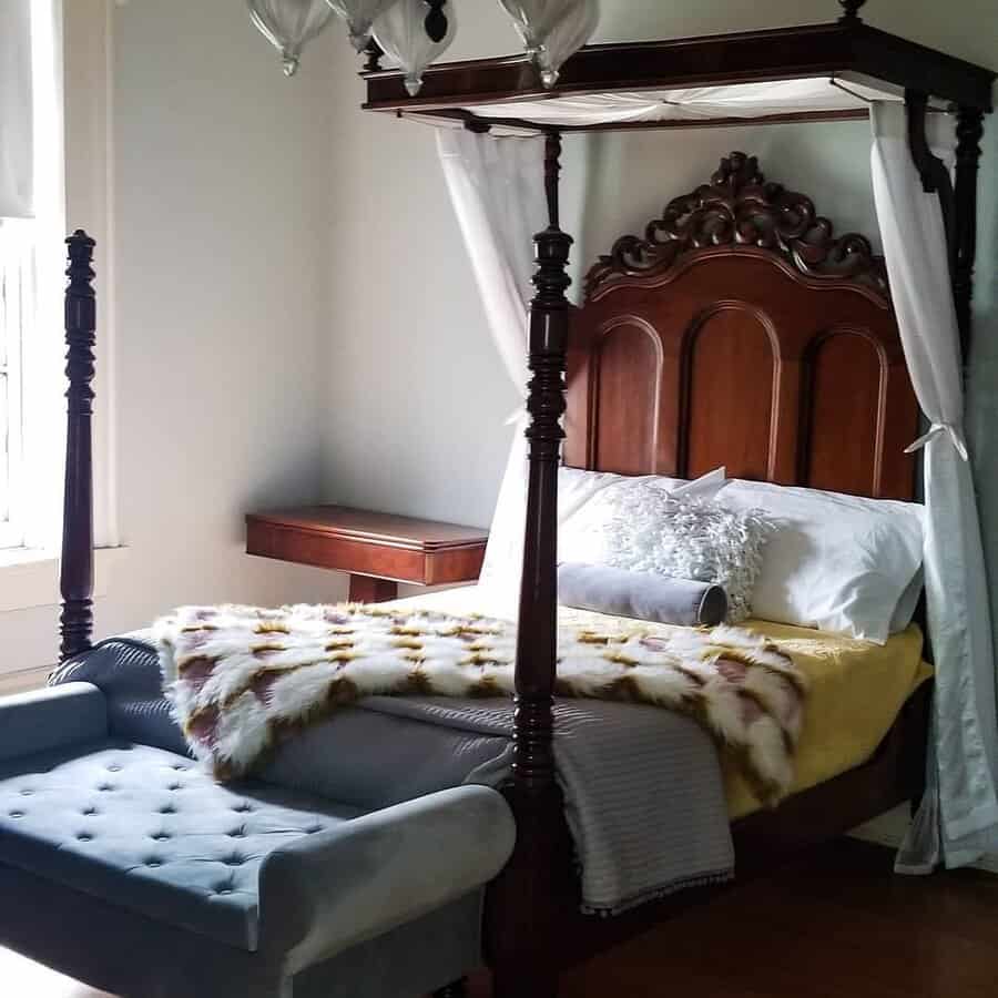 Ornate wooden canopy bed with sheer drapes, yellow and fur blankets, and tufted blue bench in a softly lit room