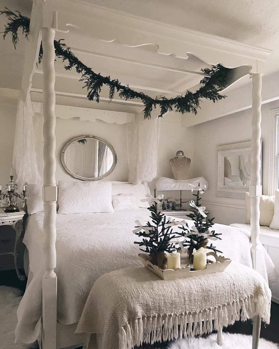 Cozy bedroom with a white canopy bed, draped greenery, and festive decor; a tray with candles and a small tree is on a bench