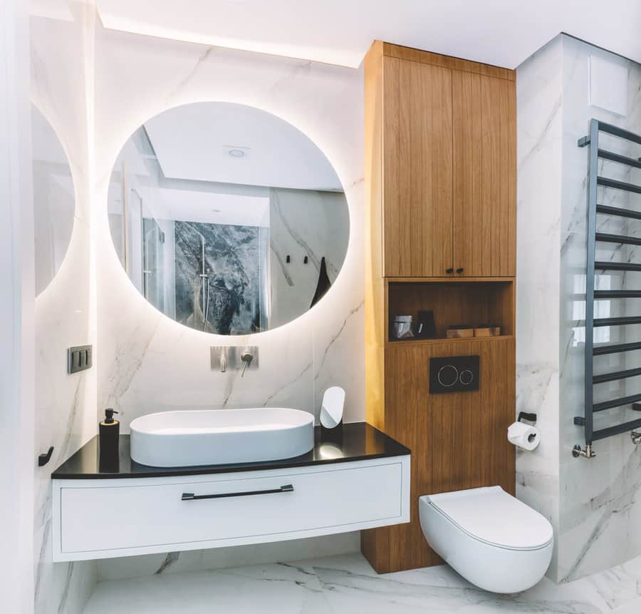 Modern bathroom with a round mirror, floating vanity, wooden cabinet, wall-mounted toilet, and towel warmer; marble walls and floor