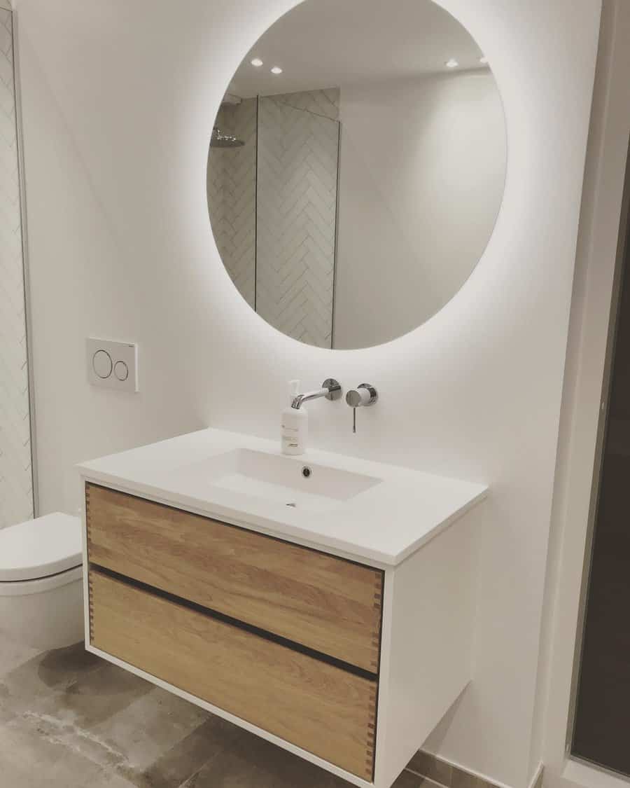 Modern bathroom with round mirror, backlit LED, wooden vanity with sink, faucet, and small soap dispenser; tiled wall in background