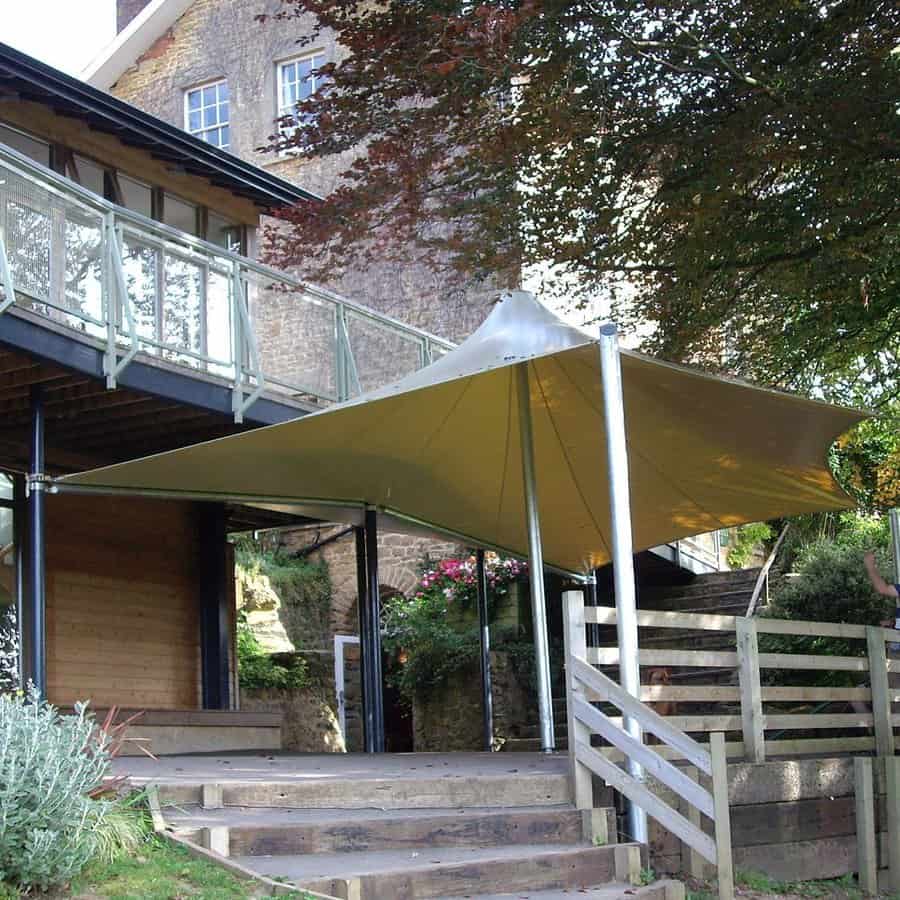 Modern free-standing shade sail with a unique peaked design, covering an outdoor patio area near a rustic stone and wood building