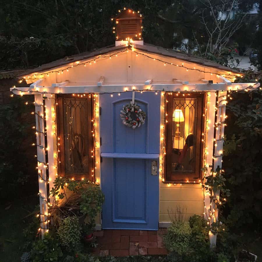 Small wooden shed with blue door, decorated with string lights and a wreath, surrounded by plants in a garden setting