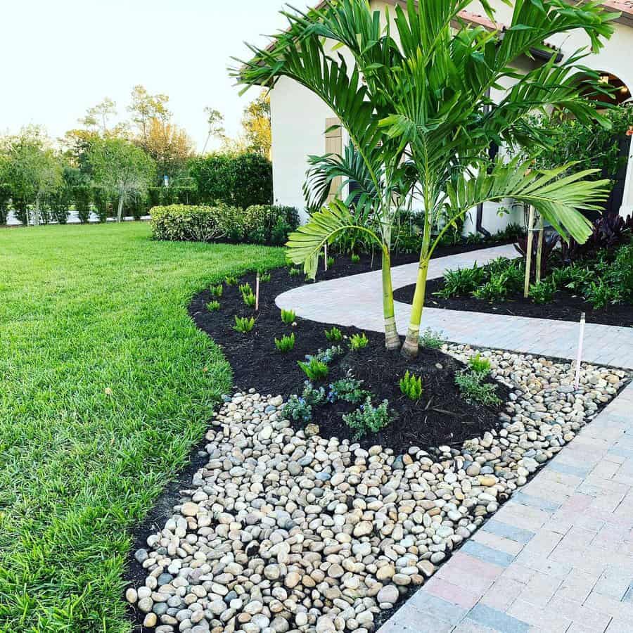 Elegant Florida front yard with palm trees, a curved brick walkway, black mulch, and decorative river rocks, creating a clean and modern tropical landscape