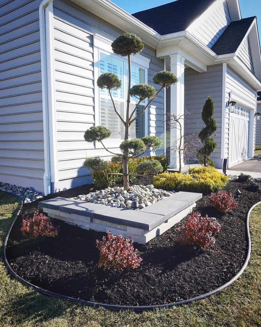 Elegant front yard landscaping with a sculpted tree on a stone pedestal, black mulch, decorative plants, and a neat border design