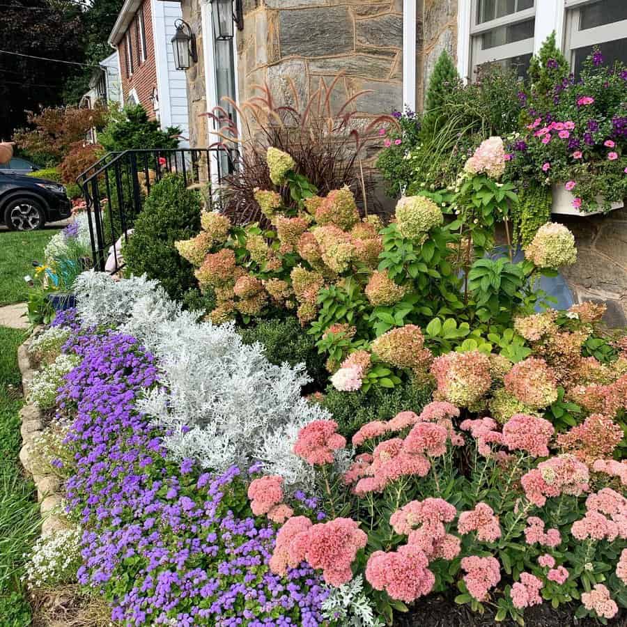 Vibrant front yard garden with hydrangeas, colorful flowers, ornamental grasses, and neatly trimmed shrubs bordering a stone house