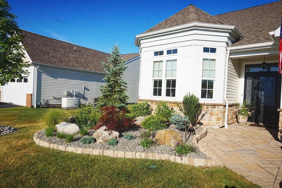 Front yard with a low-maintenance rock garden featuring evergreen trees, shrubs, decorative stones, and a paved walkway near a white house