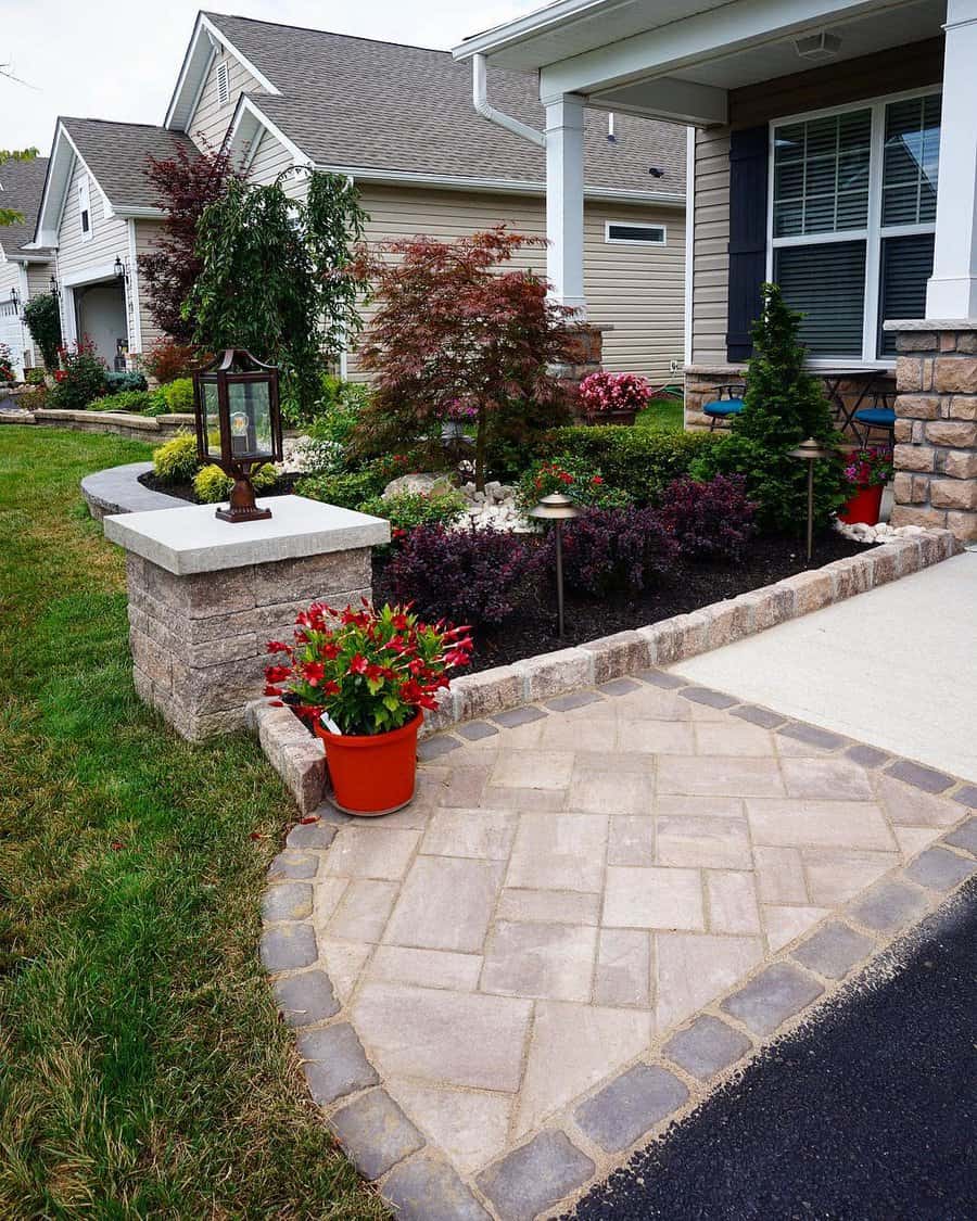 Front yard with a vibrant garden featuring a Japanese maple, potted flowers, mulch, a stone border, and a decorative paved walkway