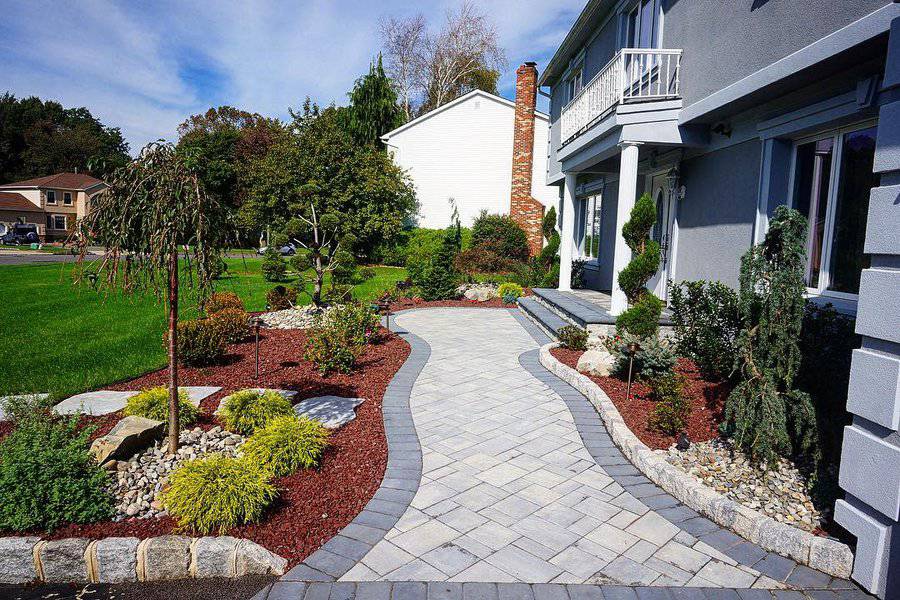 Beautiful front yard featuring a curved paver walkway bordered by vibrant mulch, decorative stones, and a variety of manicured shrubs and plants