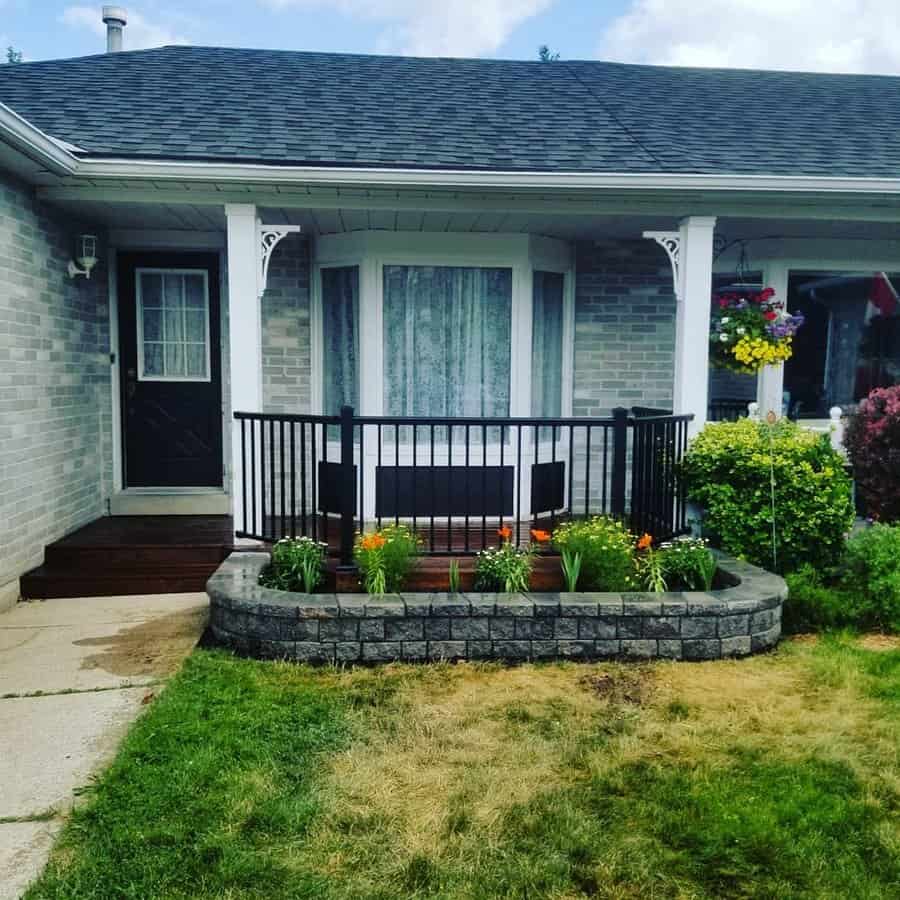 Cozy front yard landscaping with a raised flower bed, vibrant blooms, and a charming porch featuring black railing and decorative hanging baskets