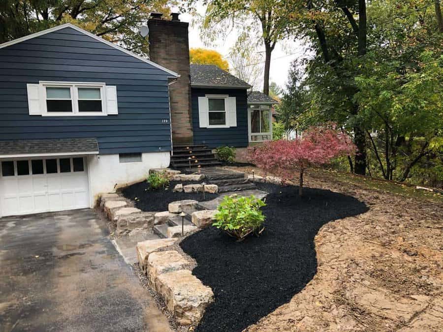 Front yard hardscaping with stone steps, black mulch landscaping, a small tree, and a navy blue house surrounded by greenery