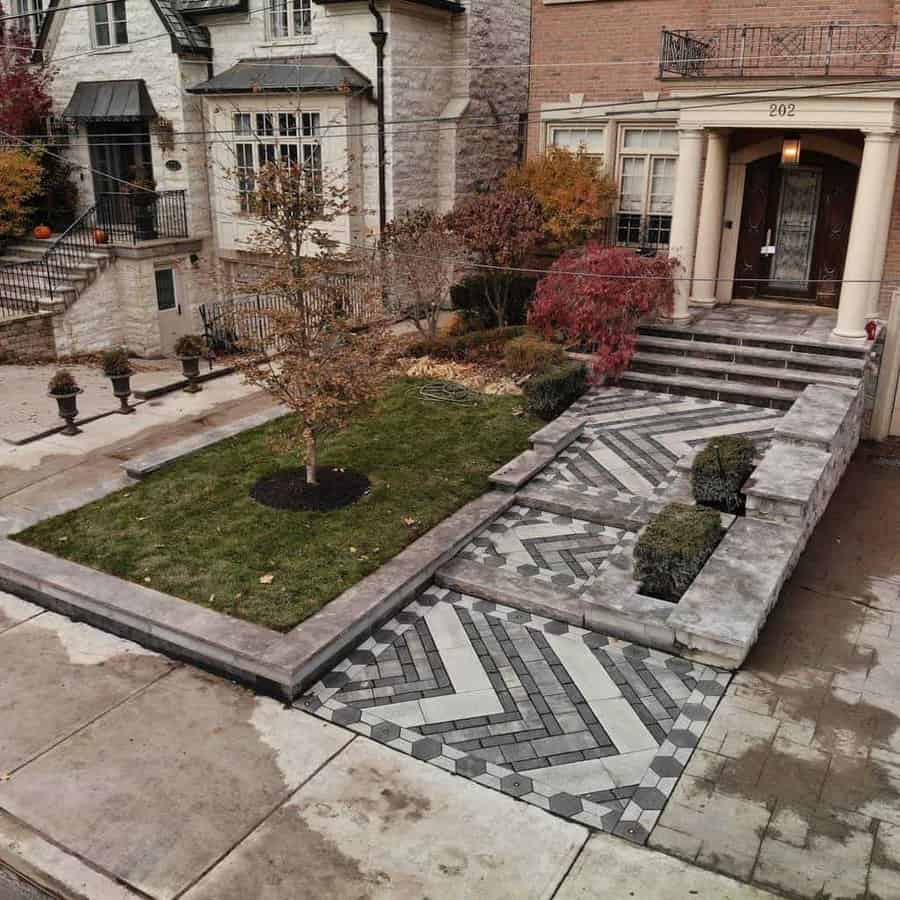 Elegant front yard with herringbone-patterned pavers, a manicured lawn, trees, and stone steps leading to a classic-style home