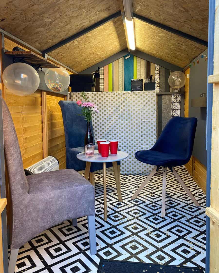 Cozy shed interior with two chairs, patterned floor, small table with a vase of flowers, red cups, balloons, and colorful back wall