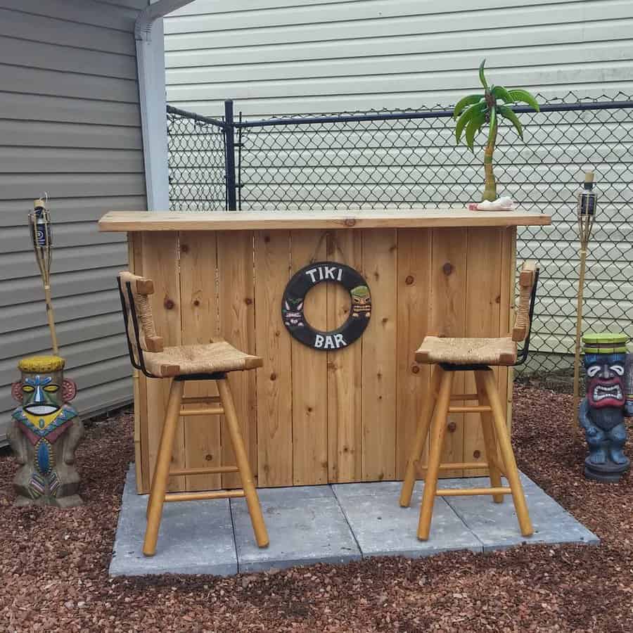 Outdoor tiki bar with two straw-topped stools, a life ring sign, tiki statues, and torches under a small roof