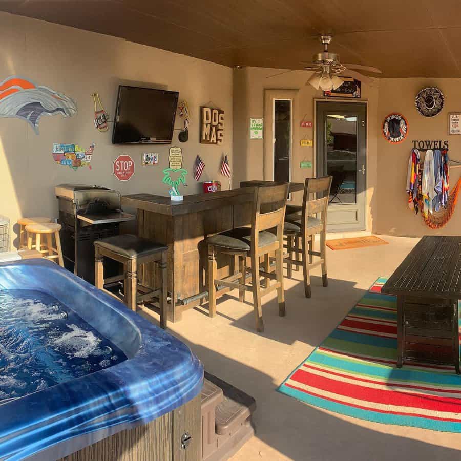 Covered patio with hot tub, bar stools, and a wall-mounted TV featuring decor with various signs and team logos along with a colorful rug on the floor