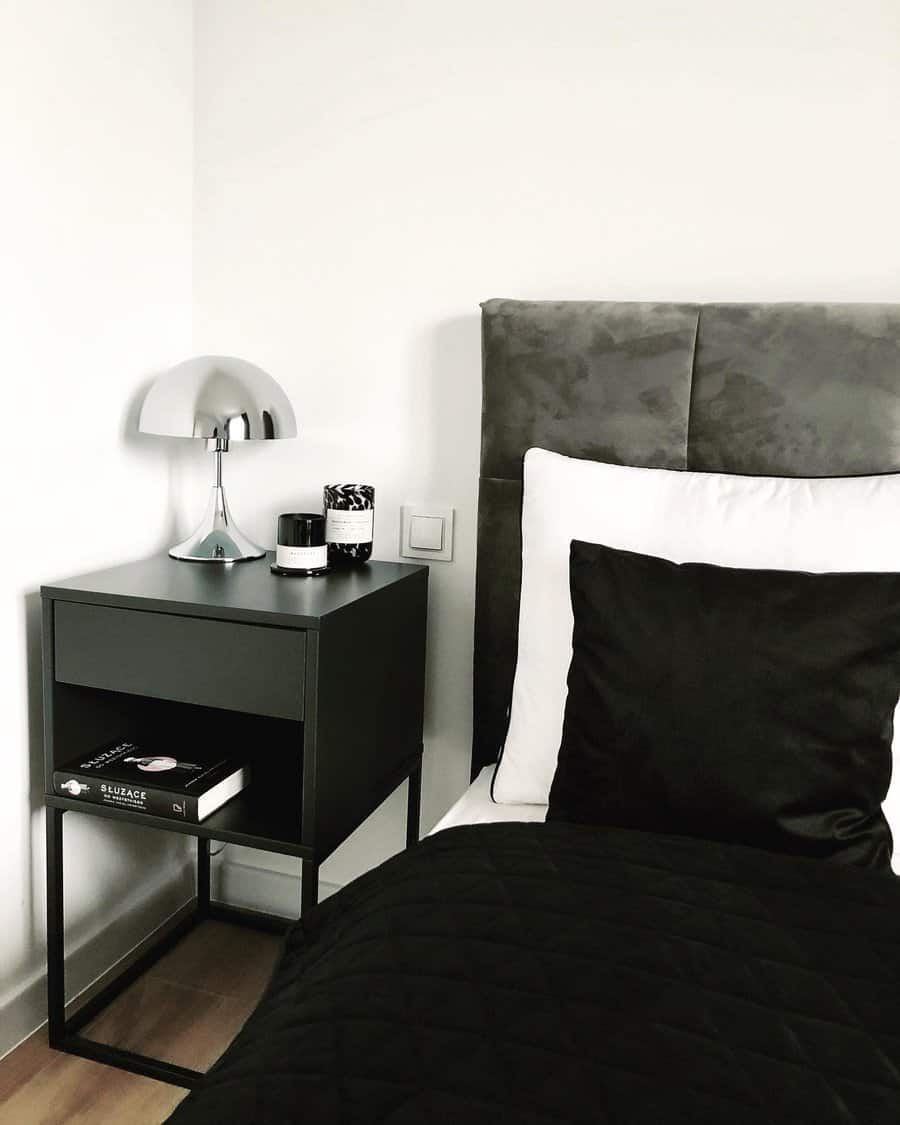 Minimalist bedroom with a gray bed, black pillow, black blanket, and a black nightstand holding a lamp, candles, and books