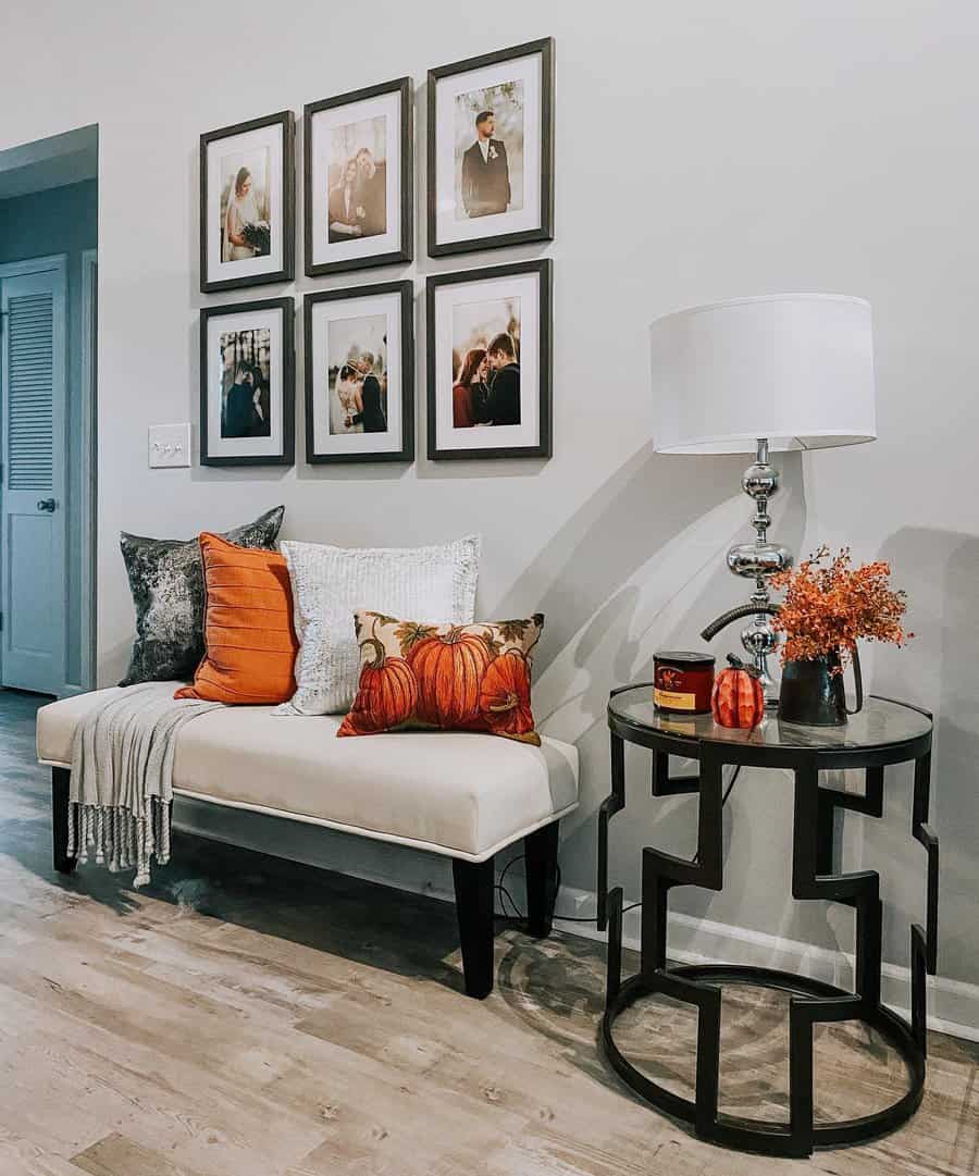 Cozy entryway with a white bench, colorful pillows, side table with lamp, autumn decor, and framed photos on a gray wall