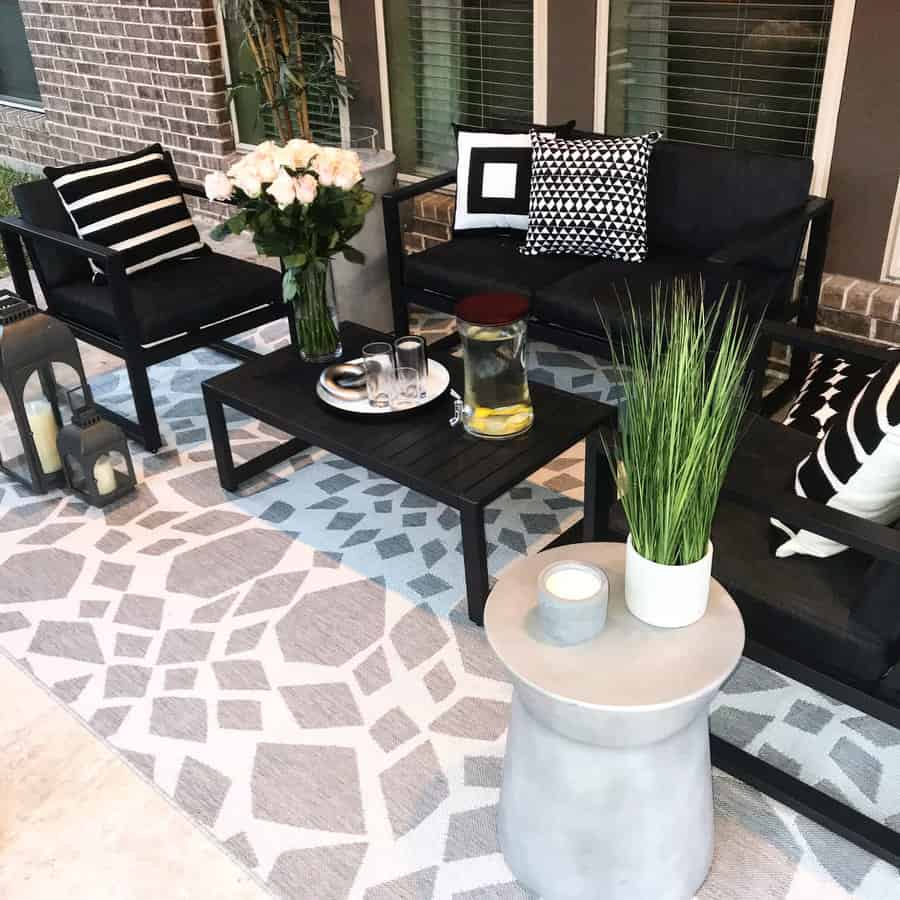 Modern patio with black furniture, patterned cushions, and a geometric rug. A table holds a pitcher, glasses, and a bouquet of flowers.