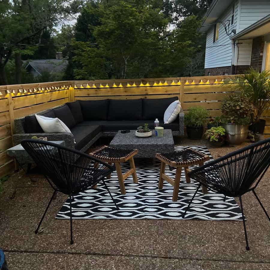 Cozy patio with wicker sofa and chairs, patterned rug, string lights, and plants; wood fence encloses the seating area