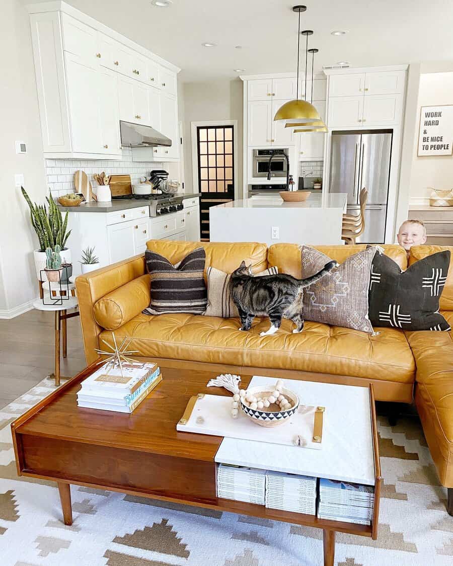 Stylish living room with a tan leather sofa, a cat on the cushions, and a smiling child peeking over; modern kitchen in the background