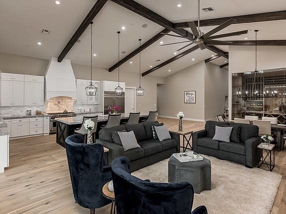 Modern open floor plan with vaulted ceilings, dark beams, plush seating, and a sleek white kitchen with pendant lighting