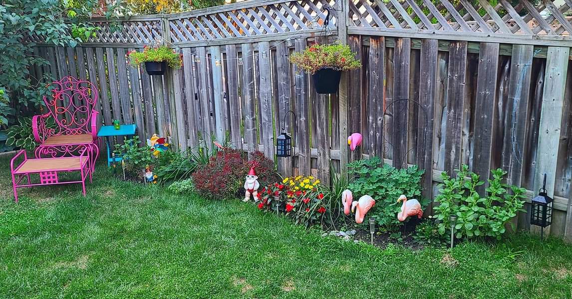 Garden with pink chair, hanging plants, flamingo statues, and flowers beside a wooden fence