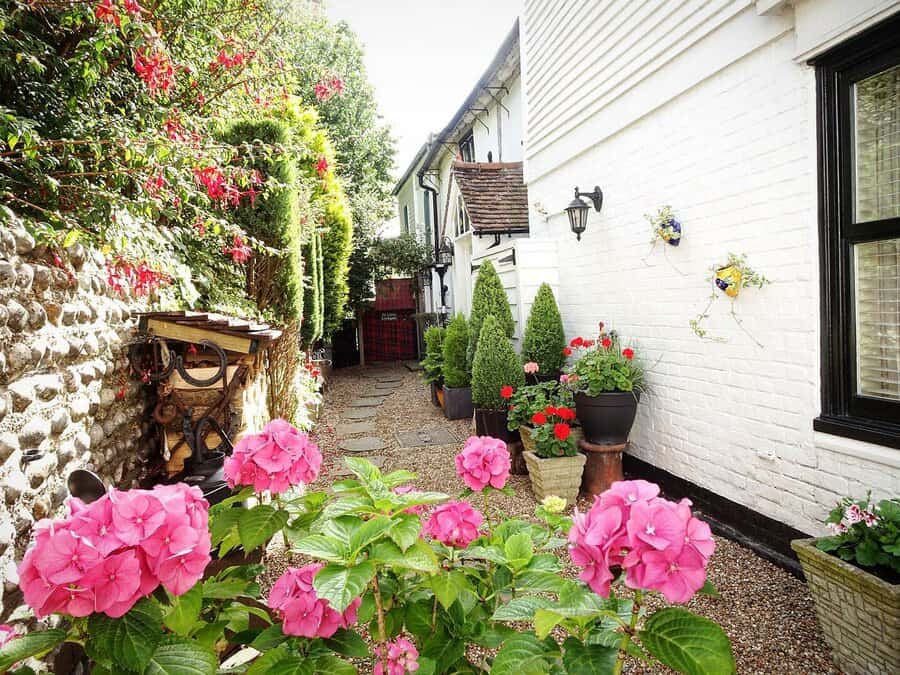 Narrow garden path with pink flowers, potted plants, and green foliage alongside white brick walls, leading to a distant gate