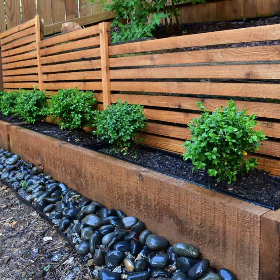 Wooden planters with small green shrubs, bordered by black stones, against a wooden slat fence in a garden setting