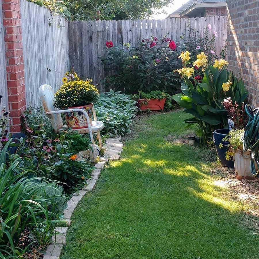 Narrow garden with green grass path, colorful flowers, potted plants, a chair, and a wooden fence under sunlight