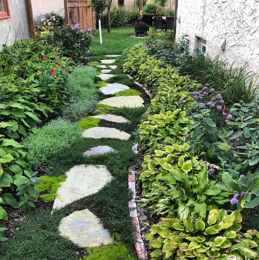 A narrow garden path with stone steps winds through lush greenery and colorful flowers between two houses