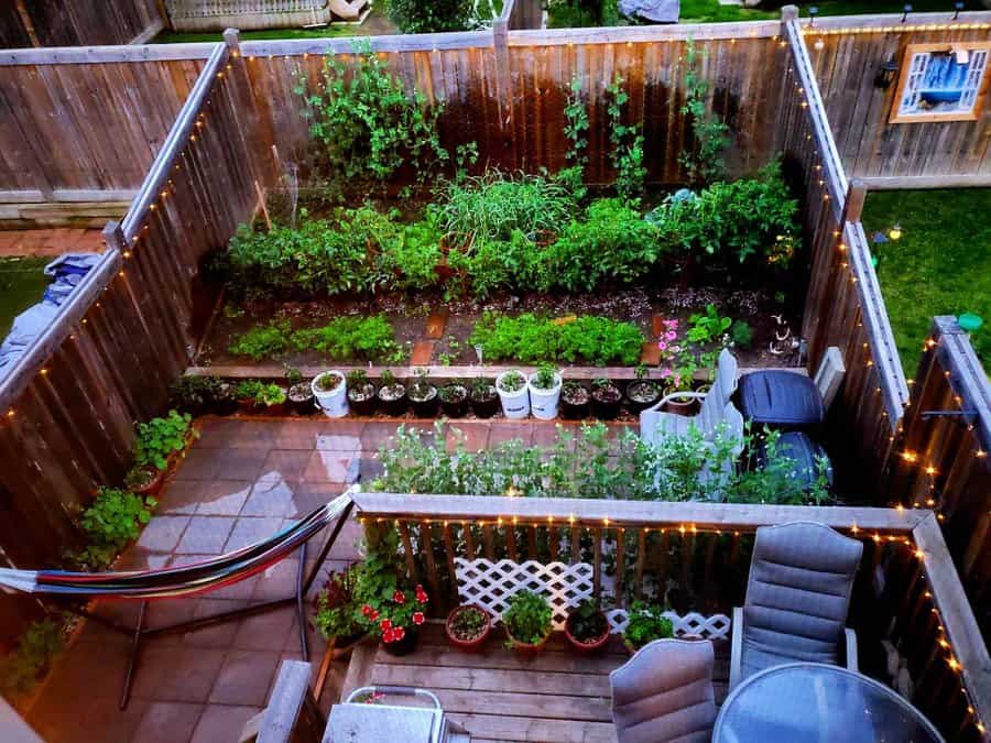 A backyard garden with raised beds, potted plants, and a hammock. String lights adorn the fence and a patio set is on the deck