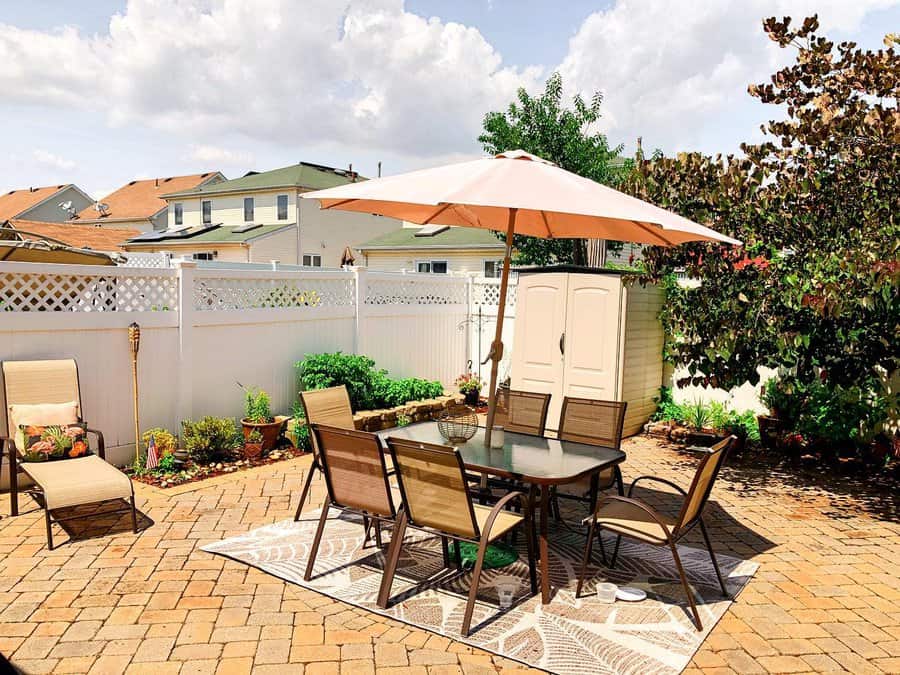 Sunny brick patio with a table, chairs, and a large umbrella on a patterned rug. Surrounded by plants, white fence, and neighboring houses