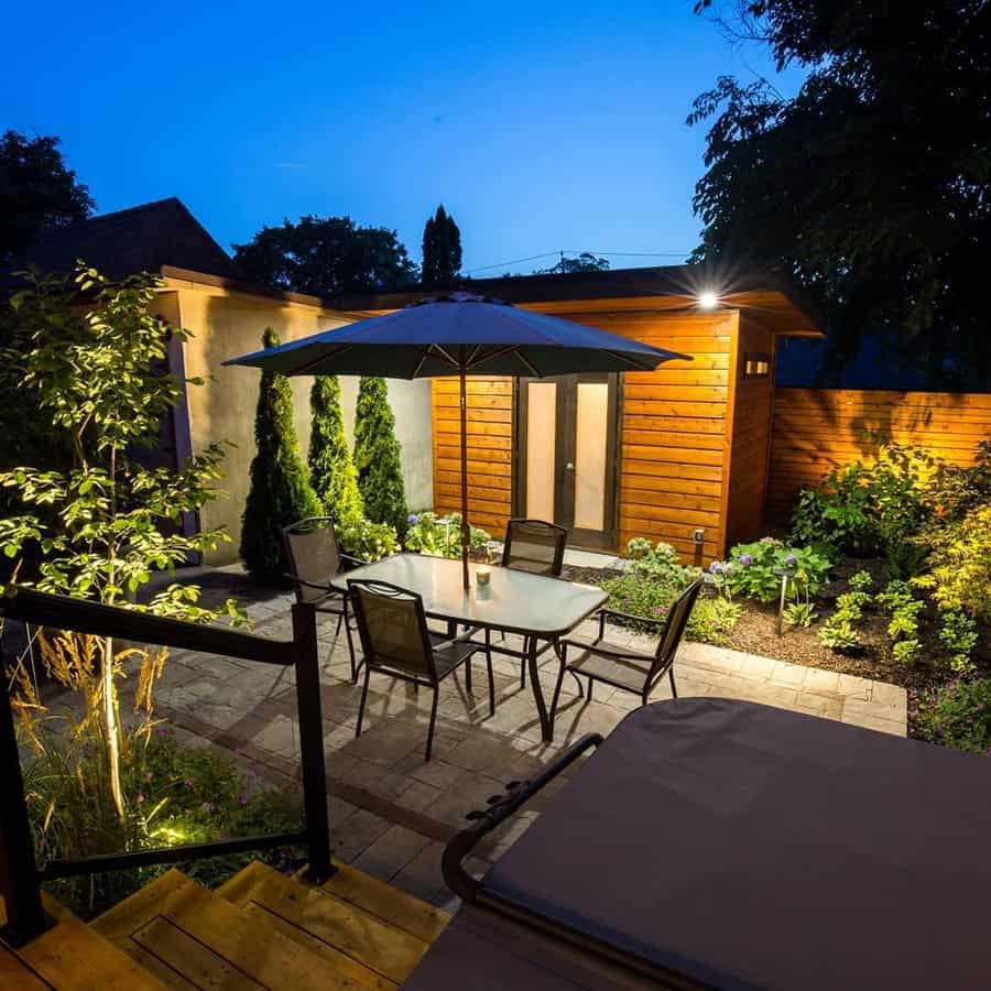 A cozy backyard with a lit patio, table, chairs, umbrella, and wooden shed, surrounded by plants, photographed at dusk