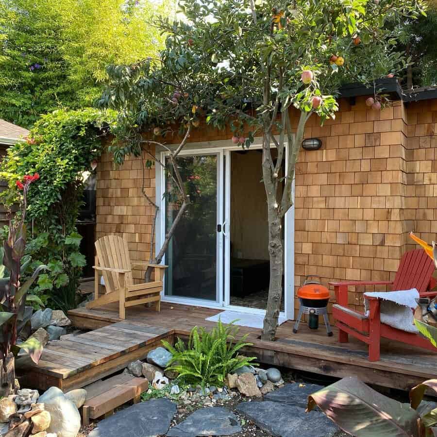 A cozy wooden cabin with a small deck, surrounded by lush greenery and trees, with two chairs and a grill in the garden area