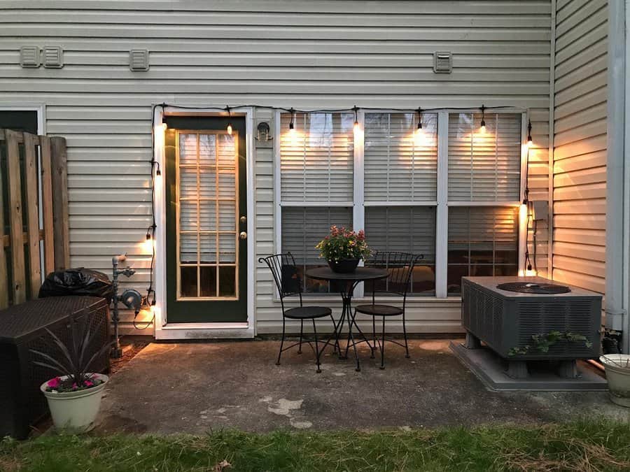 Cozy patio with string lights, a small table and chairs, potted plants, and an air conditioning unit against a white siding house