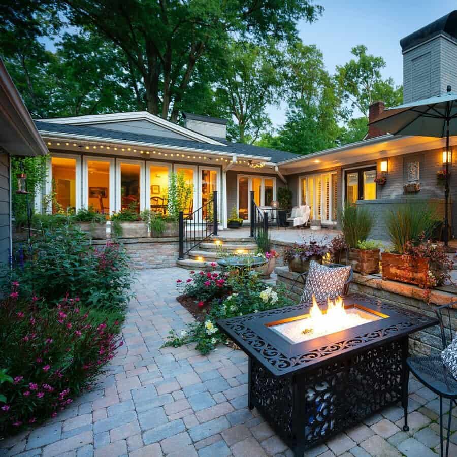 Cozy patio with a fire pit, surrounded by flowers and greenery, leading to a warmly lit house with large windows at dusk