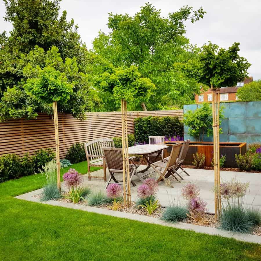 Garden with a patio set surrounded by lush greenery, trees, and ornamental grasses, with a wooden fence and blue paneling providing a backdrop