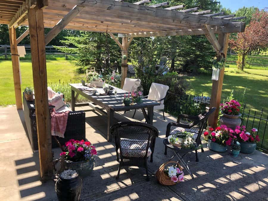 A wooden pergola with a dining table and chairs underneath, surrounded by greenery and colorful potted flowers on a sunny day